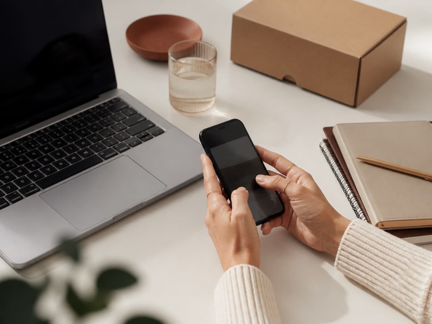Hands using a phone beside a laptop and a delivery box